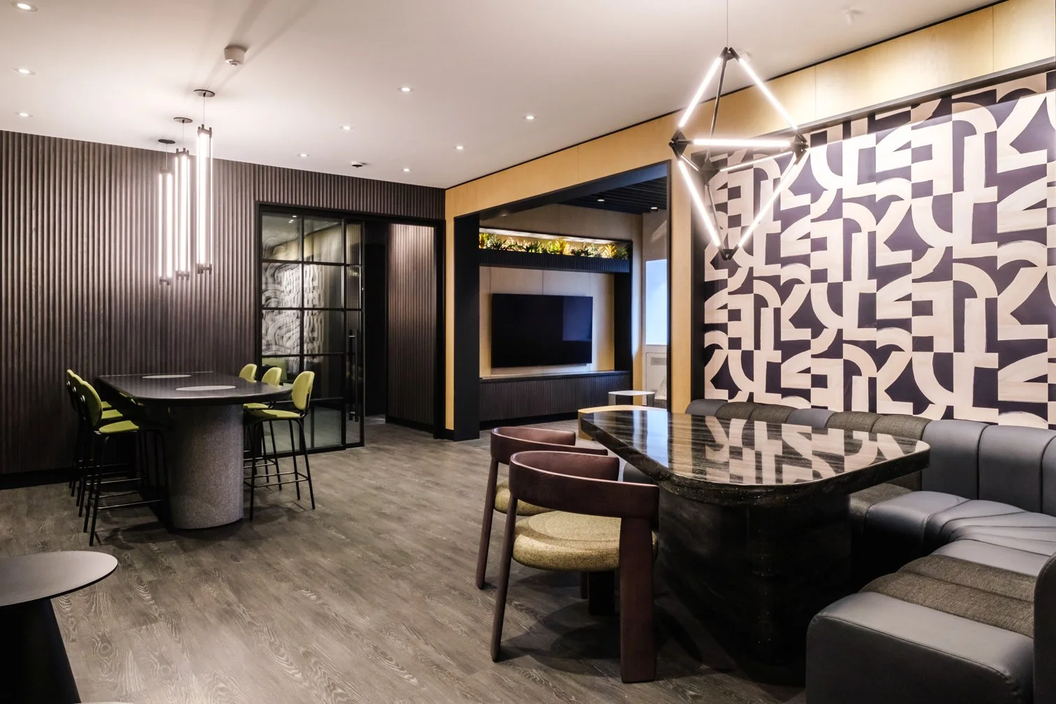 Modern interior living or lounge area with dark wood flooring, black and green chairs, a large black marble dining table, a wall-mounted TV, abstract wall art, and contemporary lighting fixtures.