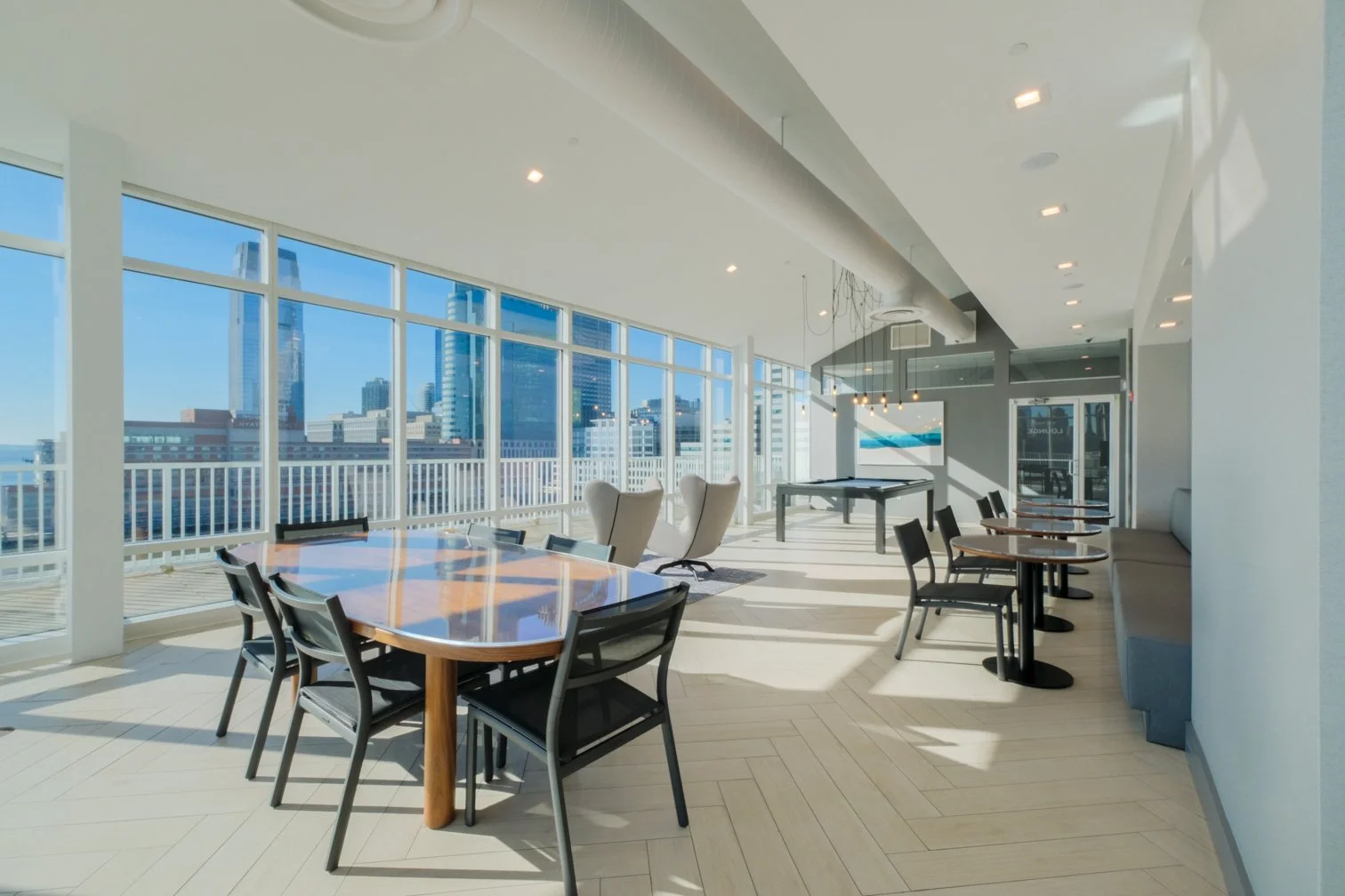 Modern lounge area with large windows showing city skyline, featuring tables and chairs, sunlight streaming in.