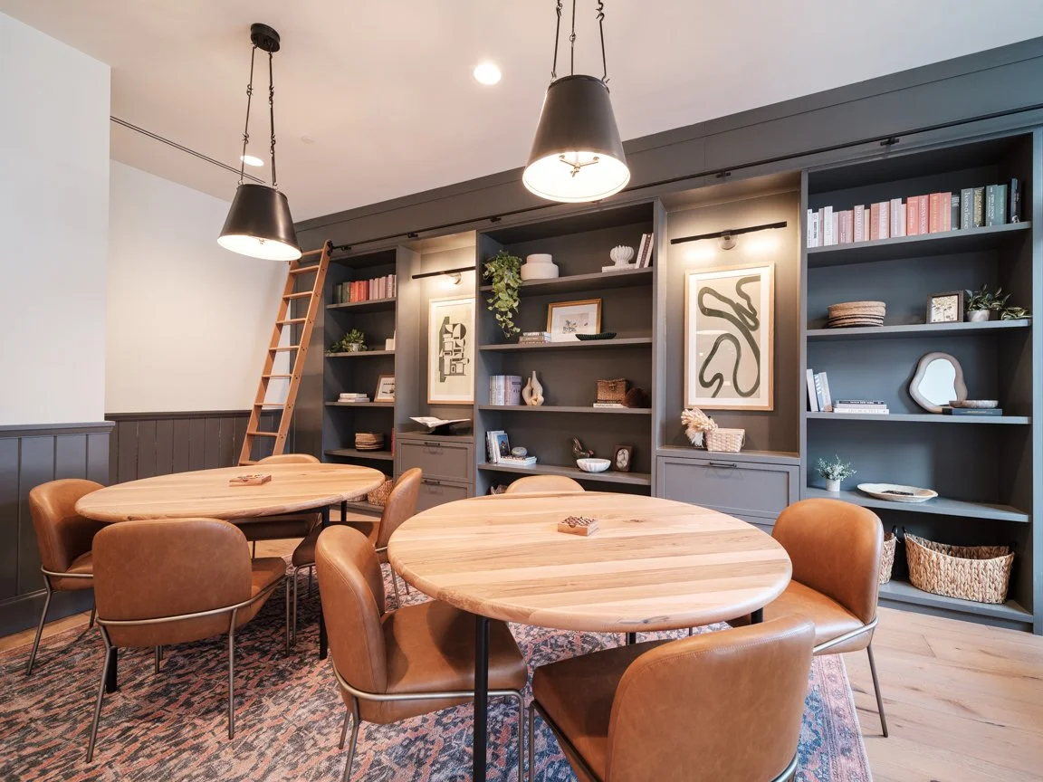 A room with two round wooden tables surrounded by brown leather chairs. A large built-in bookshelf with art, books, and decorative items is in the background, with a small wooden ladder leaning against it. Two black pendant lights hang from the ceili