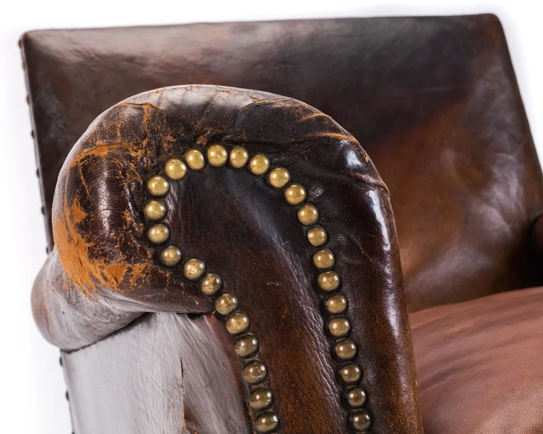 Close-up of a vintage leather armchair with worn dark brown leather and brass nailhead trim on the rolled armrest.