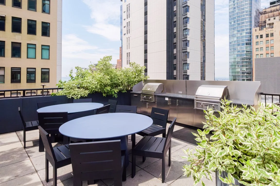 Outdoor rooftop patio with black tables and chairs, stainless steel grills, and green plants against city skyscrapers.