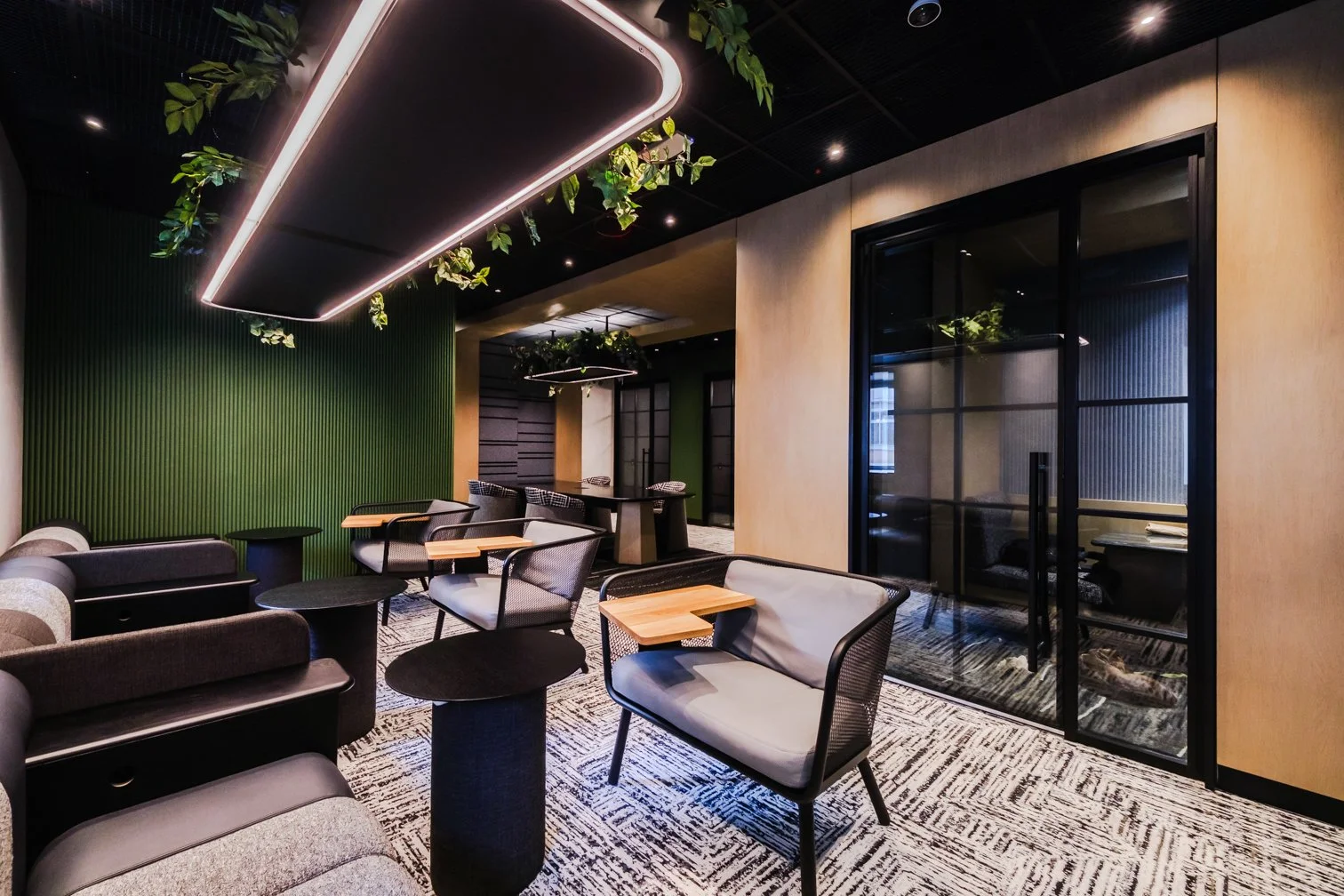 A modern lounge area with black, gray, and green decor, featuring cushioned chairs with small wooden tables, a green textured wall, black sliding glass doors, and decorative ceiling elements with hanging plants.