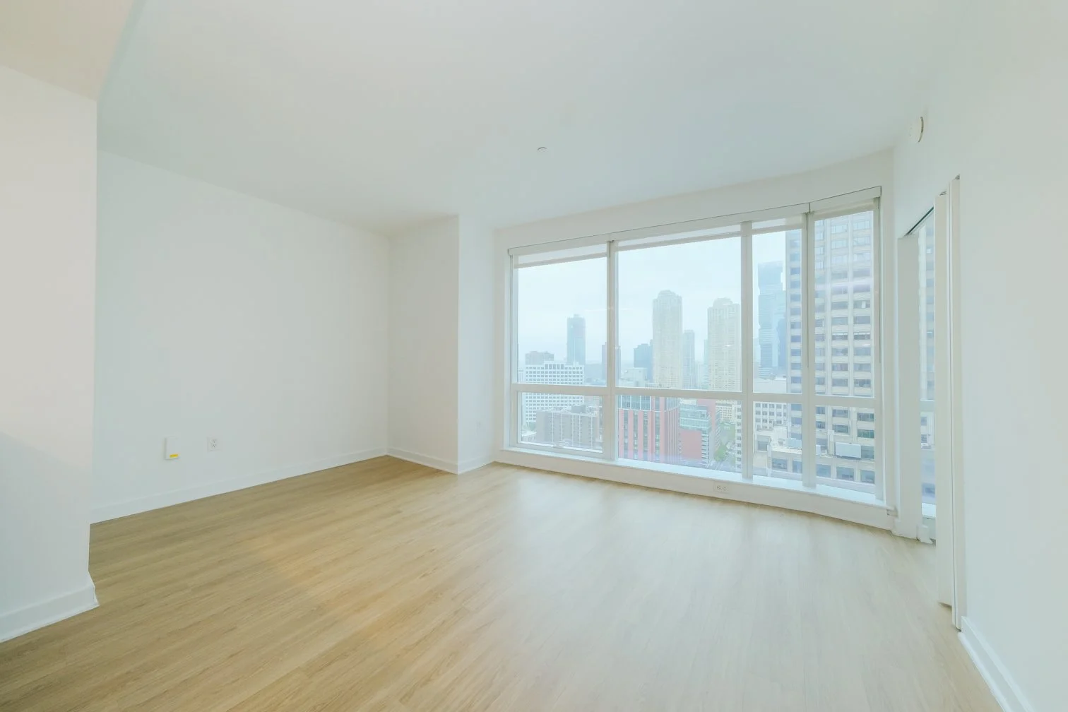 Empty room with large windows revealing a city skyline, light-colored wooden floors, and white walls.