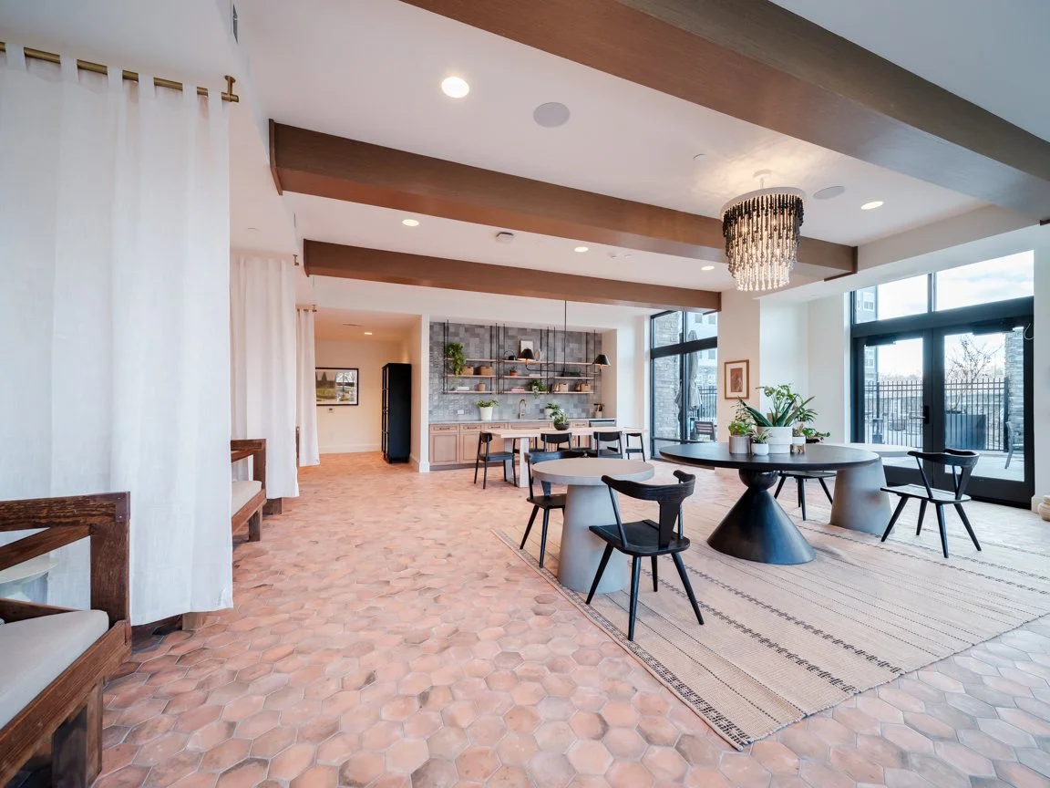 Bright, modern interior with terracotta tile floors, round dining tables with chairs, large windows, and a chandelier, featuring a minimalist decor with plants and open shelving.