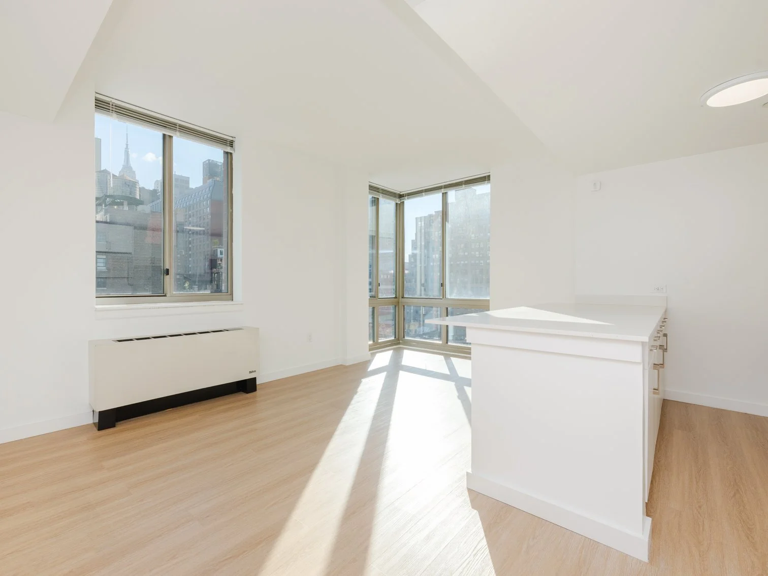Bright, empty apartment with large windows, wooden floors, white walls, and a white kitchen island.