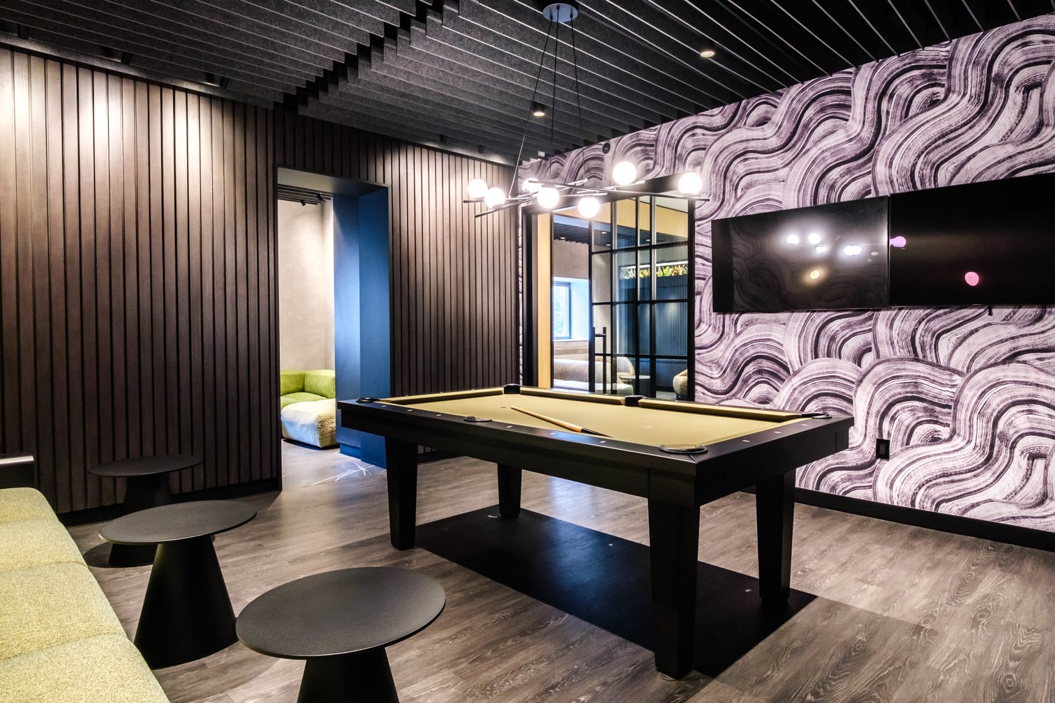 Interior of a modern game room featuring a pool table, wall-mounted TV, black furniture, textured wall, and a colorful seating area in the background.