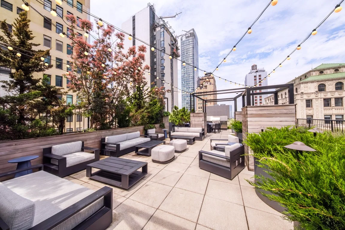 Rooftop patio with outdoor seating, string lights, trees, and city skyline in the background.