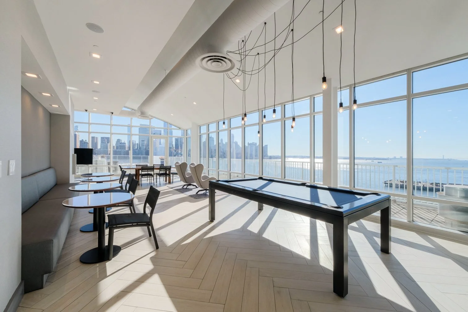 Bright interior of a modern lounge or game room with large floor-to-ceiling windows showing a city skyline and water view. Contains a pool table, seating, and hanging light bulbs.