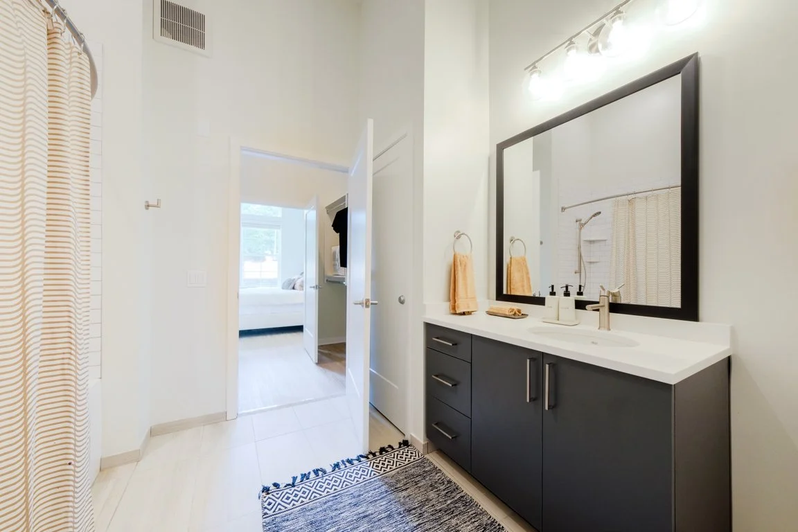Modern bathroom with dark gray vanity, white countertop, large mirror, yellow towels, and a shower curtain. Visible bedroom through open door.