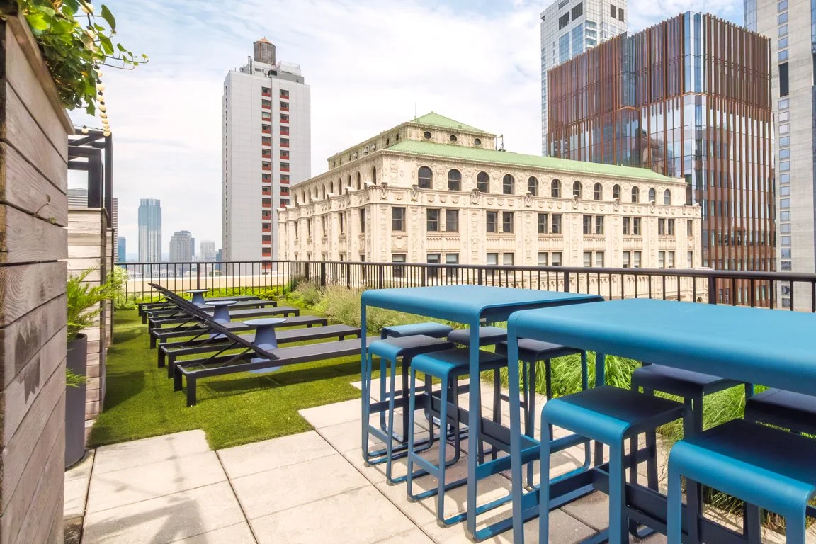 Rooftop terrace with lounge chairs and high-top tables overlooking city skyscrapers and historic building in downtown New York City.