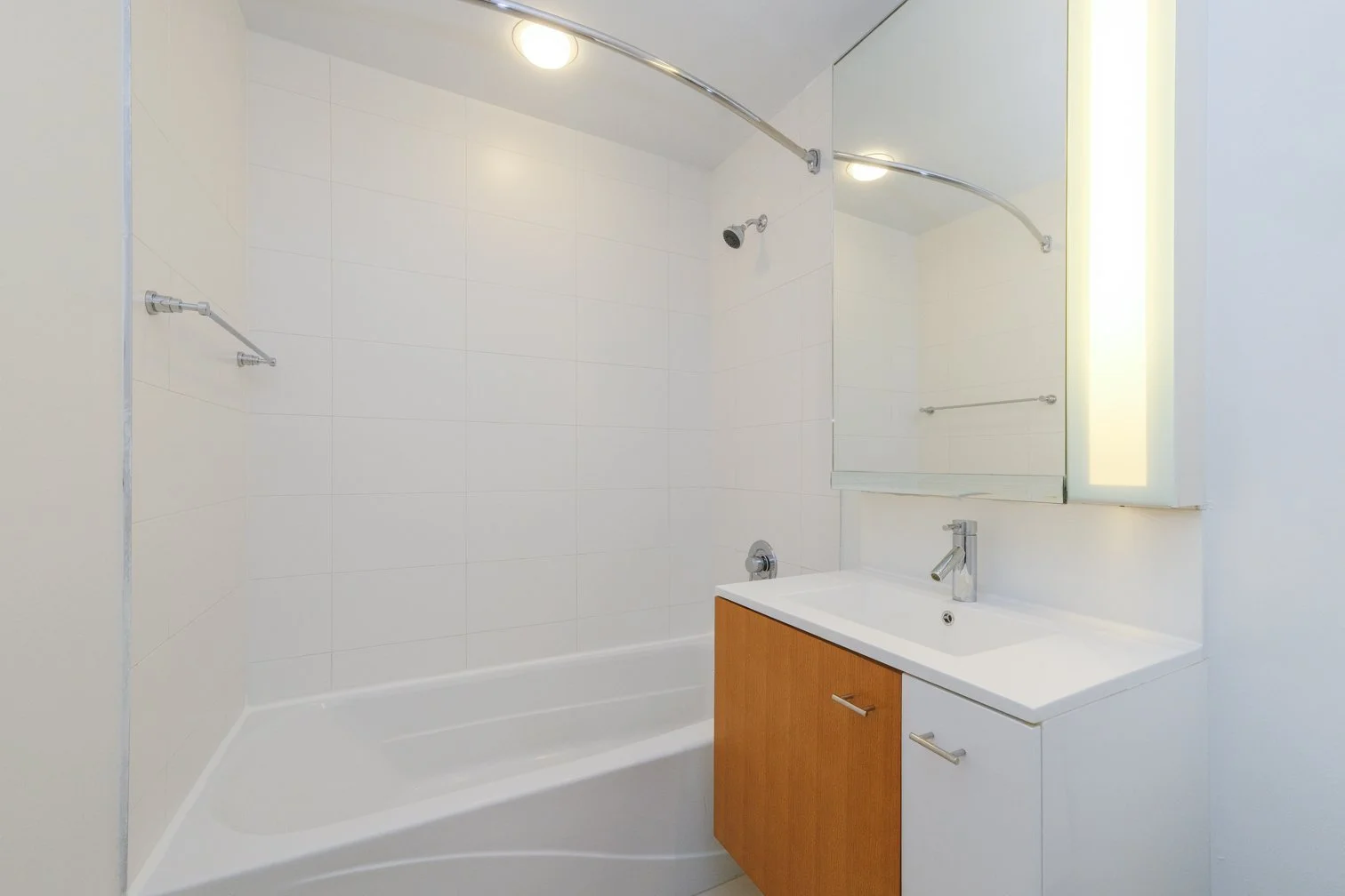 Small bathroom with a white bathtub, a wooden vanity with a sink and a faucet, a large mirror, a towel bar, and a curved shower rod with a showerhead, illuminated by ceiling lights.