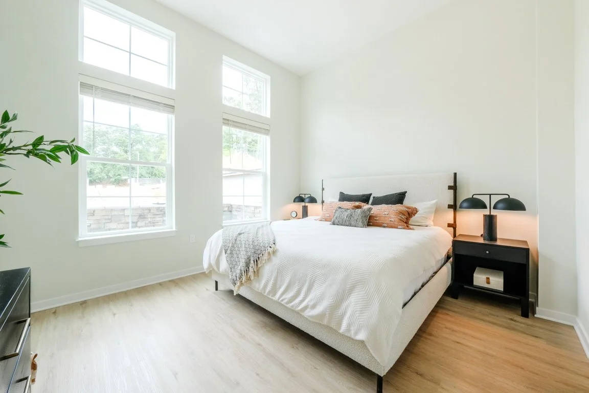 Bright bedroom with white walls, three tall windows, a white bed with gray, white, black, and brick-colored pillows, black bedside tables with black lamps, and light wood flooring.