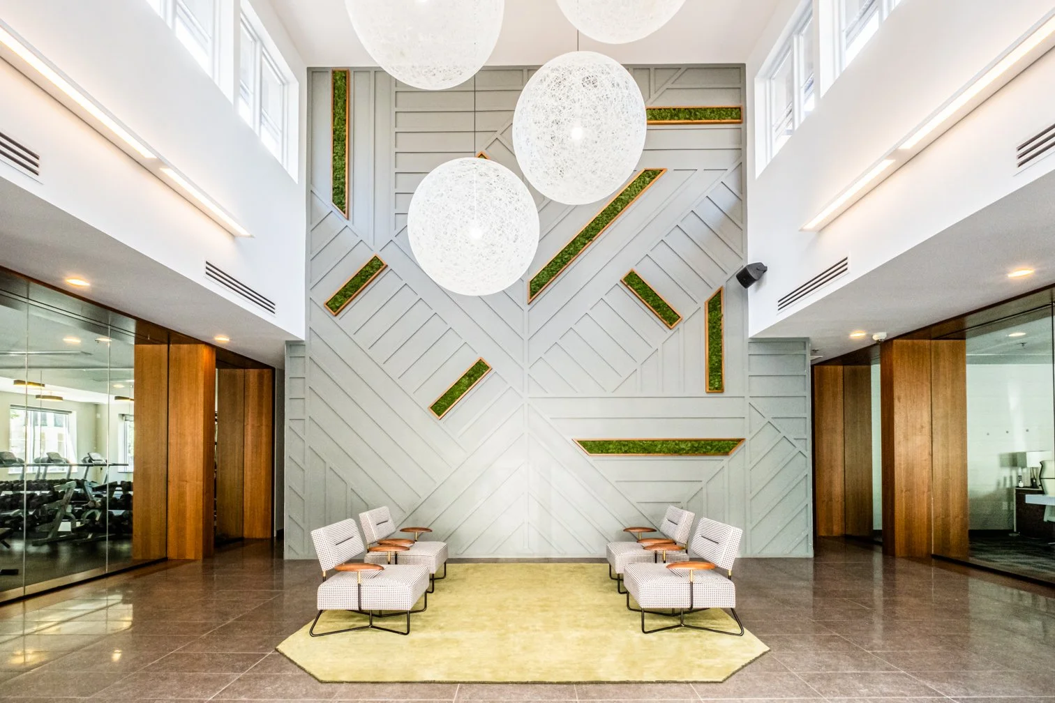 Modern lobby with white walls, large spherical pendant lights, wooden accents, a yellow rug, and seven white chairs with wooden armrests arranged in pairs on each side.