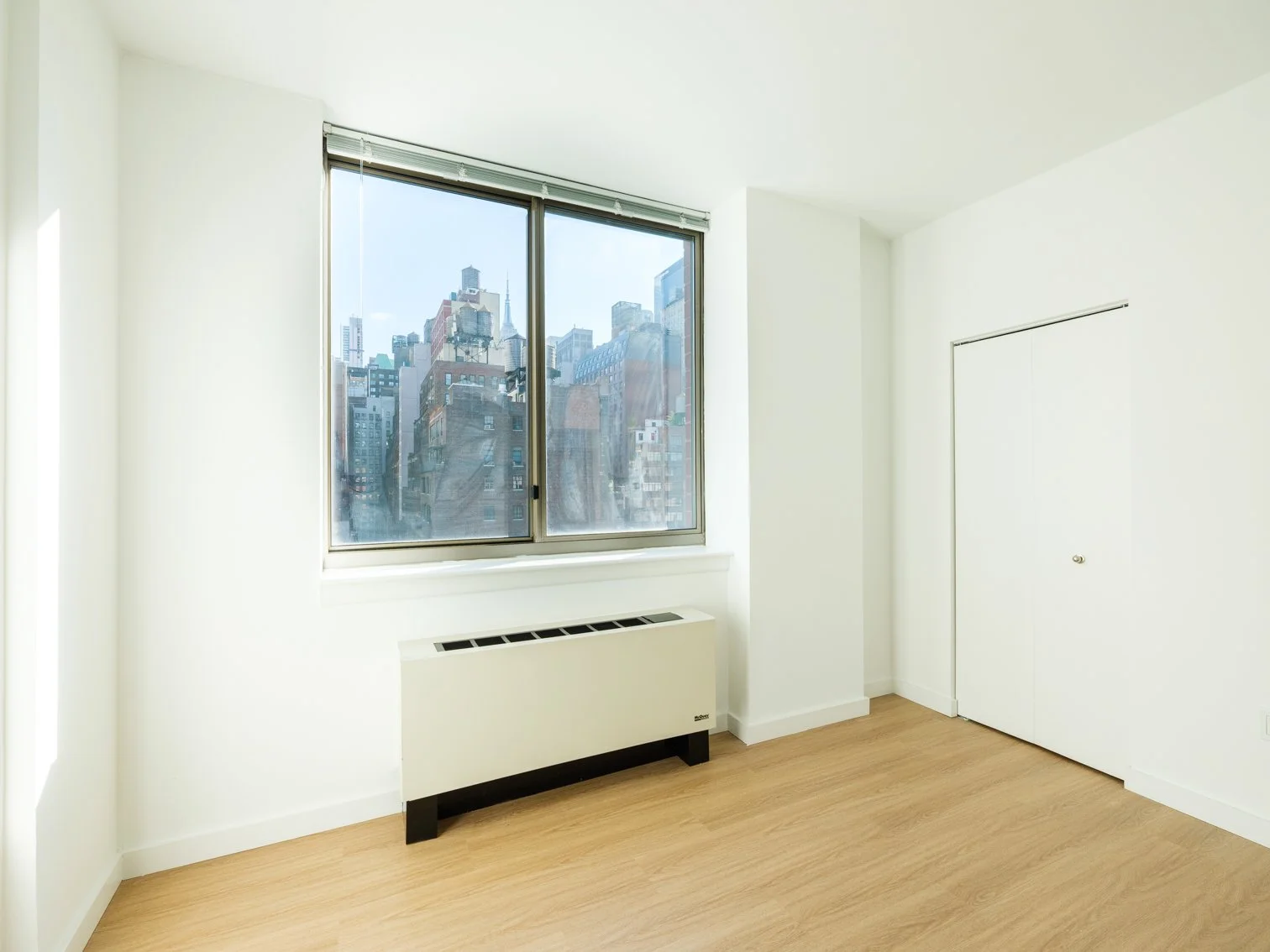 Empty room with large window showing city skyscrapers, beige floor, white walls, white closet door, and white radiator.