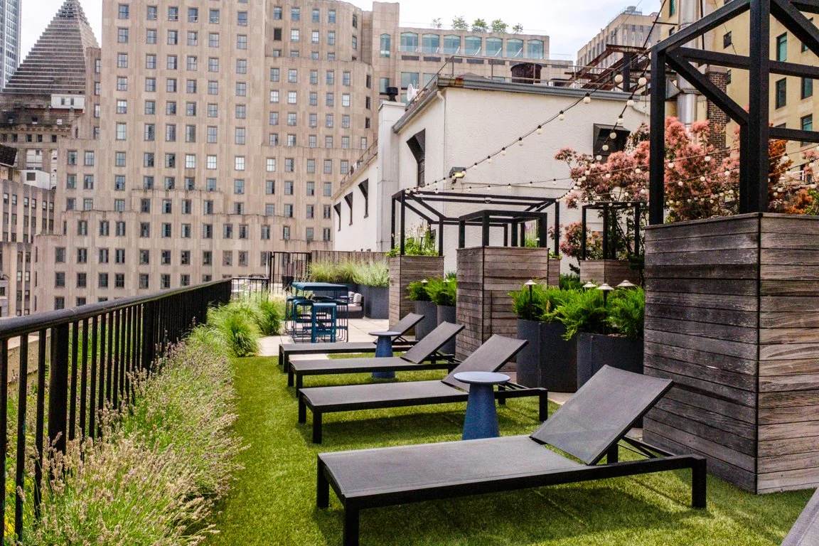 Urban rooftop terrace with lounge chairs, small tables, potted plants, string lights, surrounded by tall city buildings.