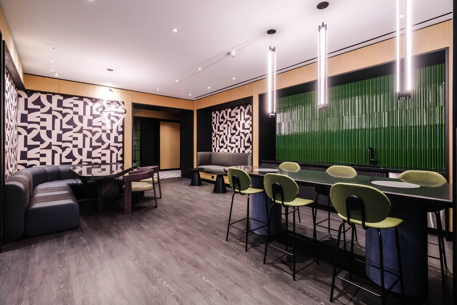 Modern living room with black, green, and beige accents, featuring a bar area with green tiled wall, multiple green bar stools, abstract wall art, a sectional sofa, and a dining table with chairs.