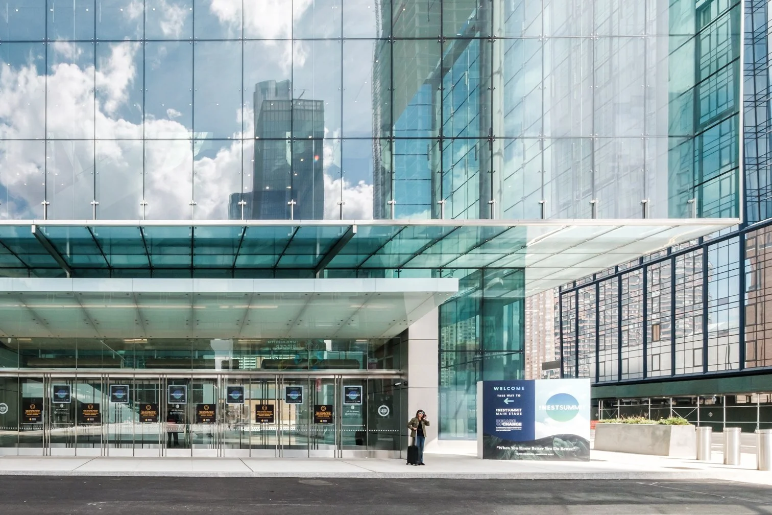 Modern glass building with reflections of clouds and neighboring skyscrapers, entrance doors, a woman with a suitcase, and signage near the entrance.