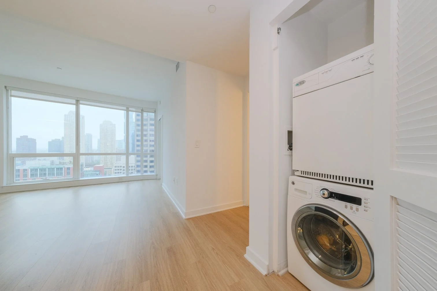 Interior view of a modern apartment with large windows overlooking a city skyline, and a laundry area with stacked washer and dryer units.