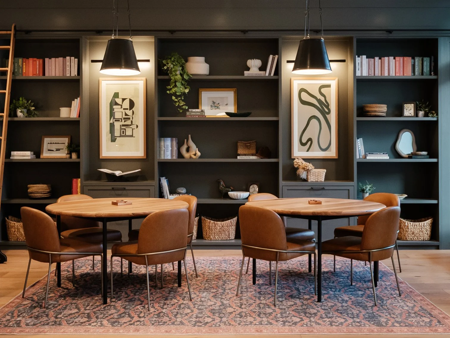 A cozy dining room with two round wooden tables, six leather chairs, a patterned area rug, and built-in dark shelving filled with books, decorative items, and art pieces, illuminated by two pendant lights.