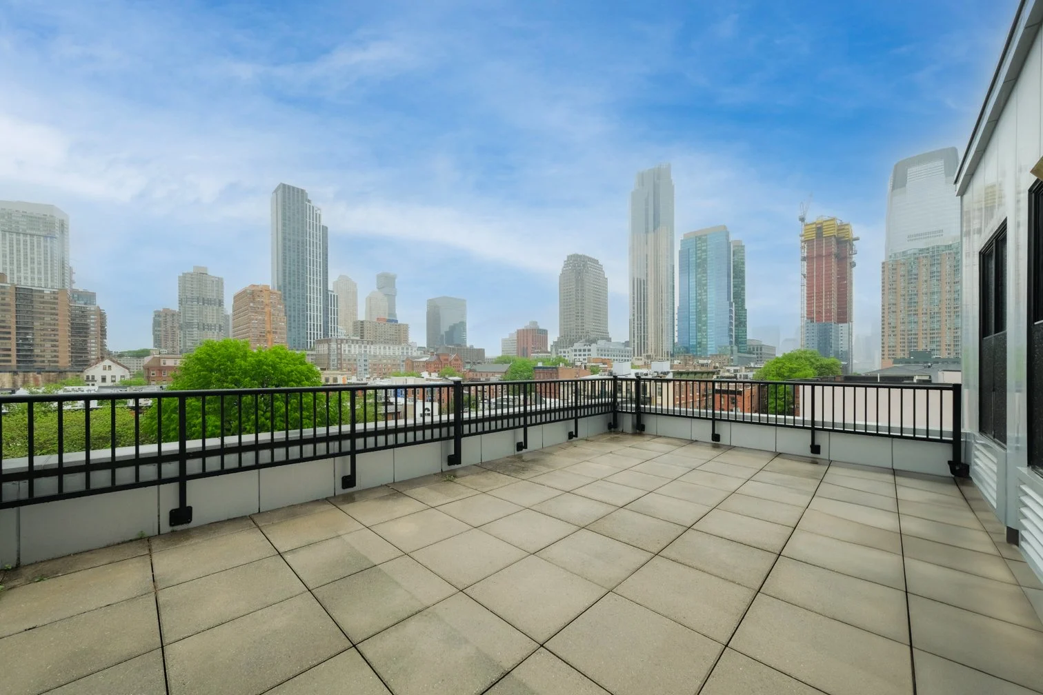 Empty balcony with black railing overlooking a city skyline with tall buildings, some under construction, and a few green trees, under a partly cloudy blue sky.