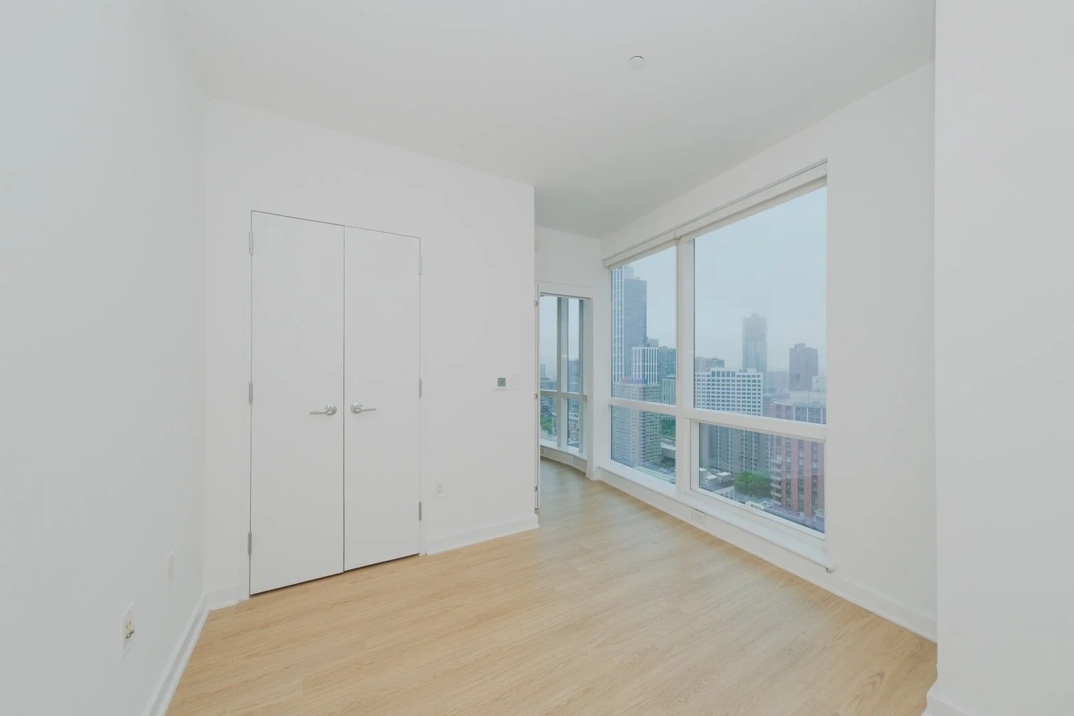 Empty room with white walls, wooden flooring, large windows with cityscape view, white double door closet, and a small balcony door.