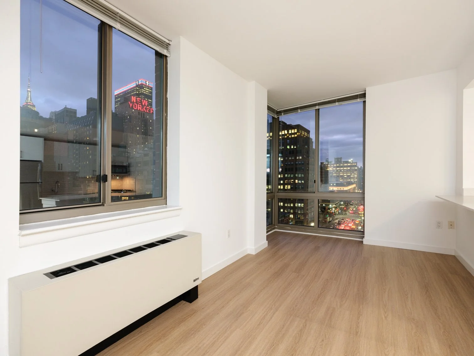 Empty room in an apartment with large windows showing cityscape at dusk, hardwood floors, white walls, and a baseboard heater.