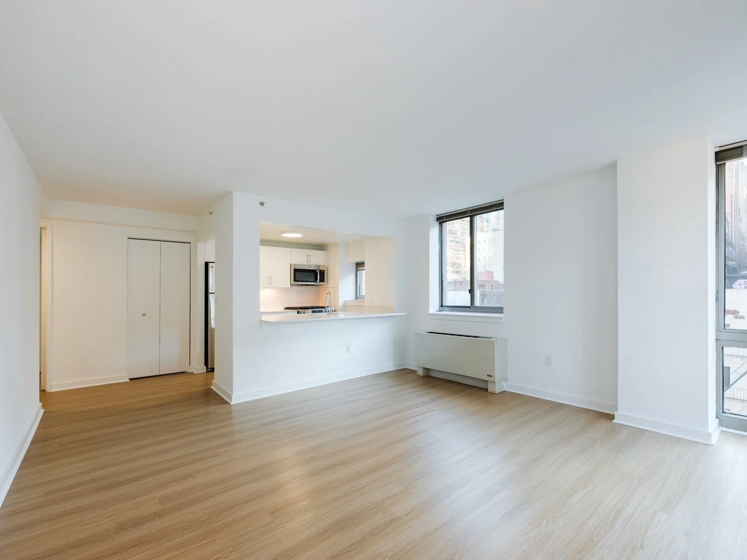 Empty living room with hardwood floors, white walls, large windows, and an open kitchen area.