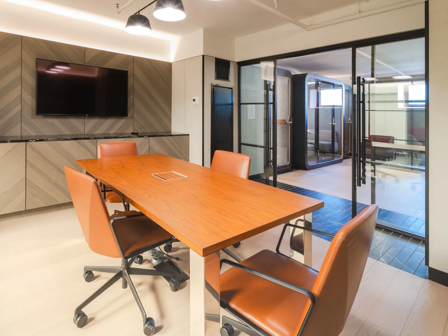 Modern conference room with a wooden table surrounded by orange leather chairs, a wall-mounted TV, and a glass wall leading to a small outdoor patio or break area.