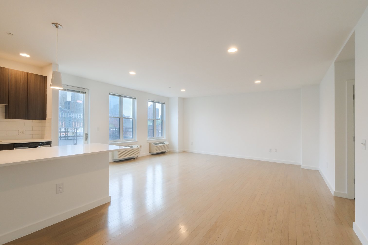 Empty living room with large windows, hardwood floors, and white walls, with a partial view of the kitchen area.
