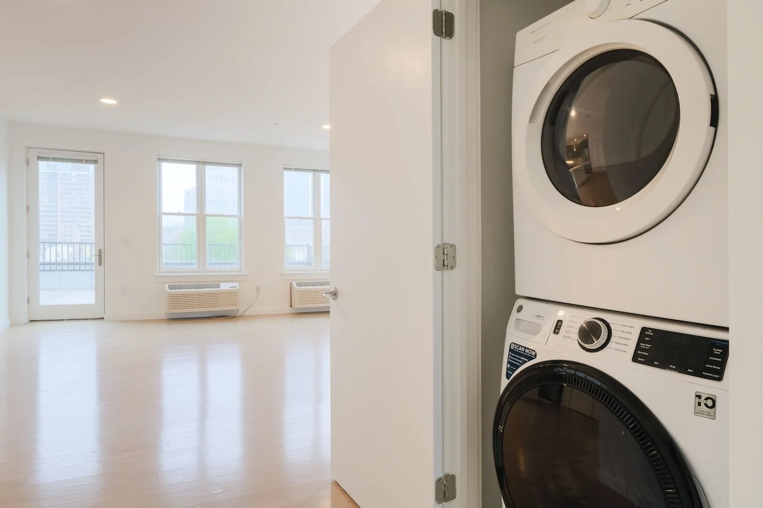Empty living room with hardwood floors, three large windows, and a balcony door, with a stacked washer and dryer in a nearby closet.