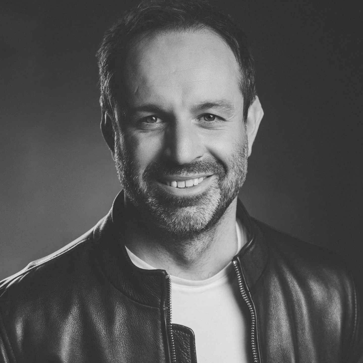 Black and white portrait of a smiling man wearing a leather jacket and white shirt.