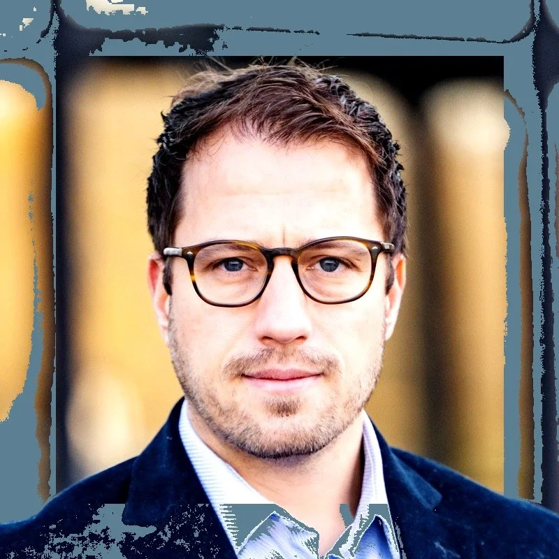 A man with short brown hair, glasses, and light skin wearing a dark blazer and a collared shirt, standing outdoors with a blurred background.