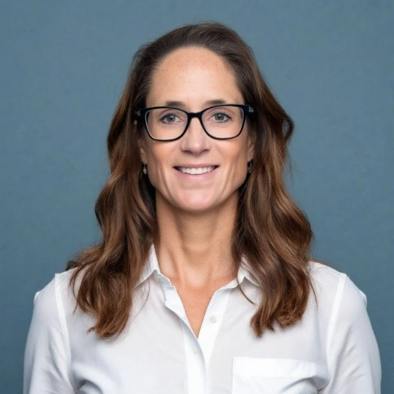 A woman with wavy brown hair and glasses smiling at the camera, wearing a white button-up shirt, against a blue background.