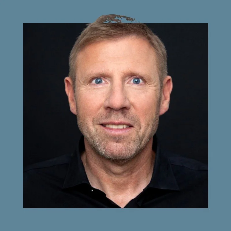 Headshot of middle-aged man with short blond hair, blue eyes, and light beard, wearing a black shirt, smiling against a dark background.
