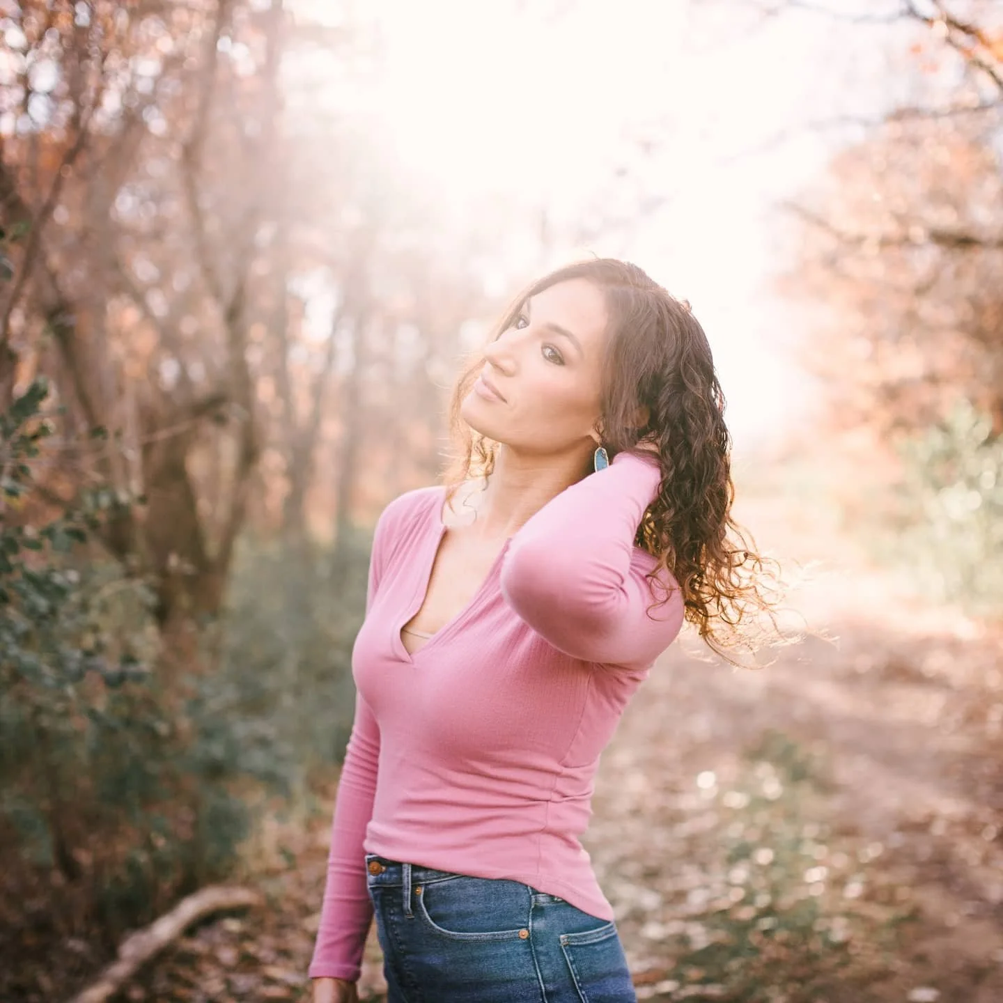 A woman with curly hair wearing a pink long-sleeve shirt and jeans standing outdoors in a sunlit forest with autumn foliage.