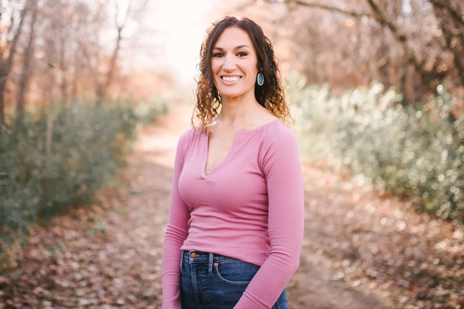 A woman smiling outdoors on a dirt path, wearing a pink long-sleeve shirt, jeans, and turquoise earrings, with trees and autumn foliage in the background.