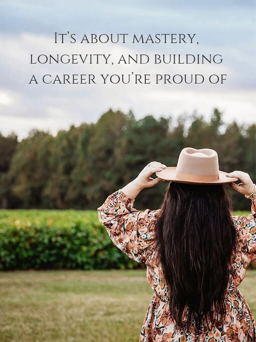 A woman with long dark hair wearing a floral dress and a beige hat standing outdoors in a grassy field with trees in the background, holding her hat with both hands. Overlaid text reads, 'It's about mastery, longevity, and building a career you're proud of.'