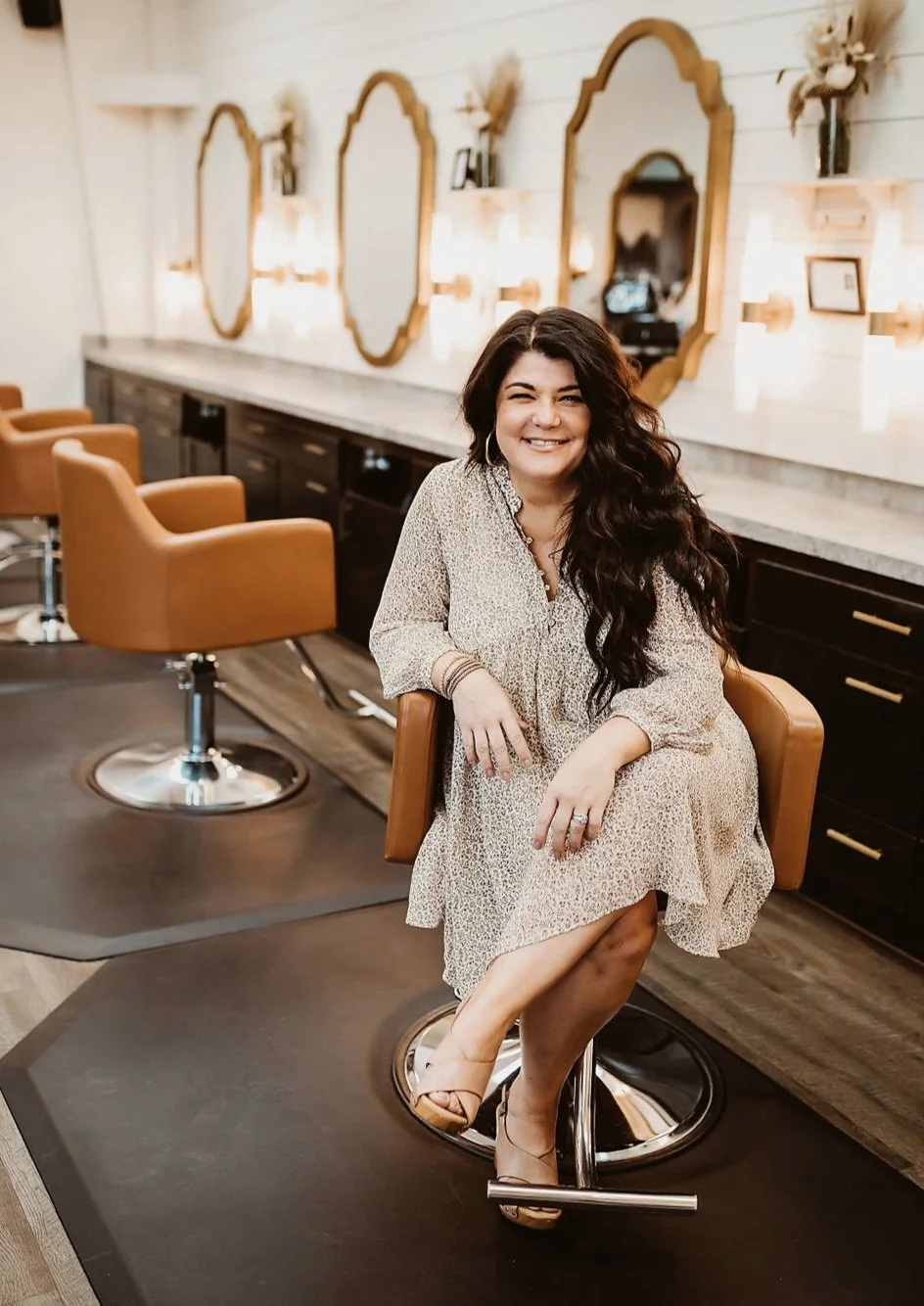 A Luna Method extension coach with long dark, wavy hair smiling and sitting on a salon chair inside a stylish hair salon with mirrors, framed pictures, and wall-mounted lamps.