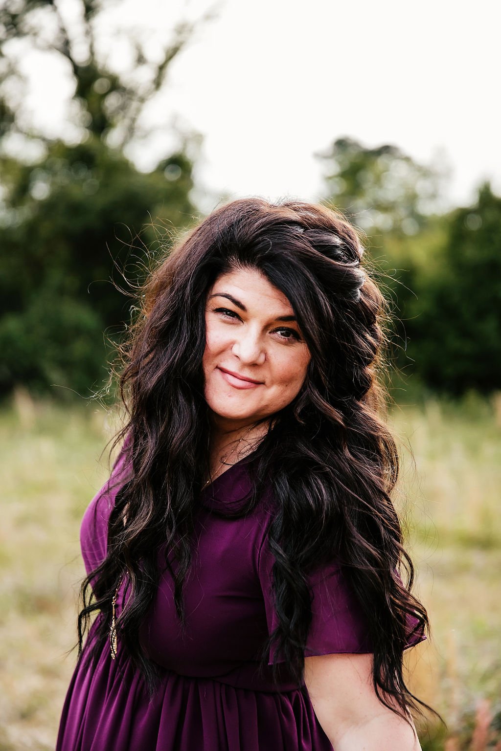 Barbi Lee, a woman with long, wavy dark hair outdoors, wearing a purple dress, smiling at the camera with a blurred background of trees and grass.