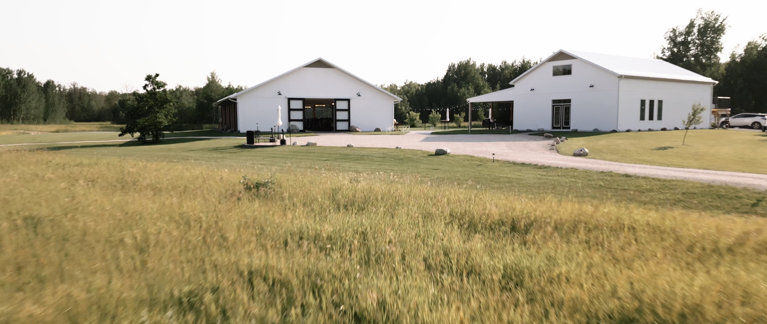 A rural property with two white buildings, a gravel driveway, and a grassy field in the foreground. The buildings have large doors, windows, and outdoor seating areas. There are trees, a car, and a clear sky in the background.