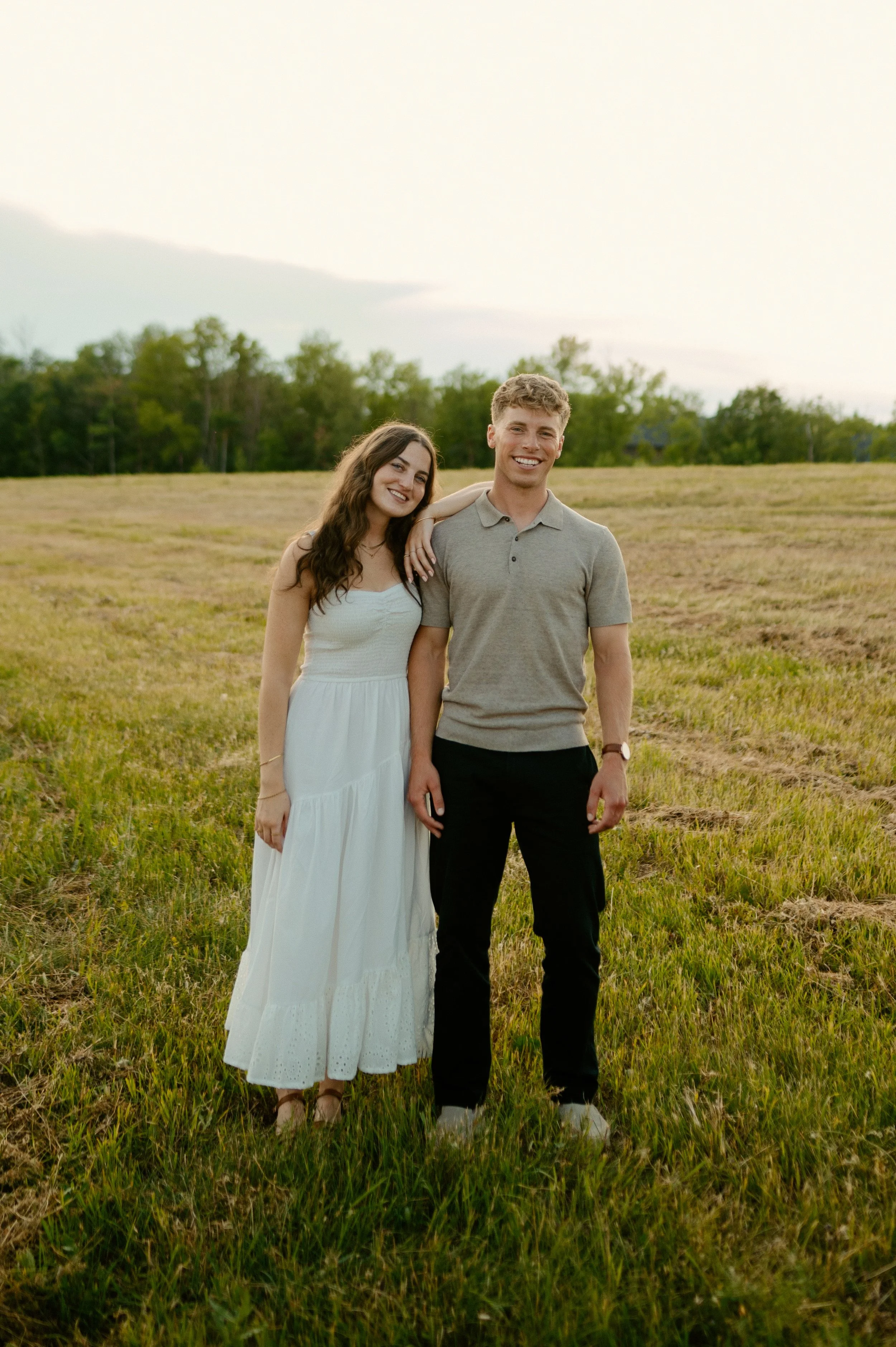 A young woman in a white dress and a young man in a gray polo shirt and black pants standing together in a grassy field during sunset.