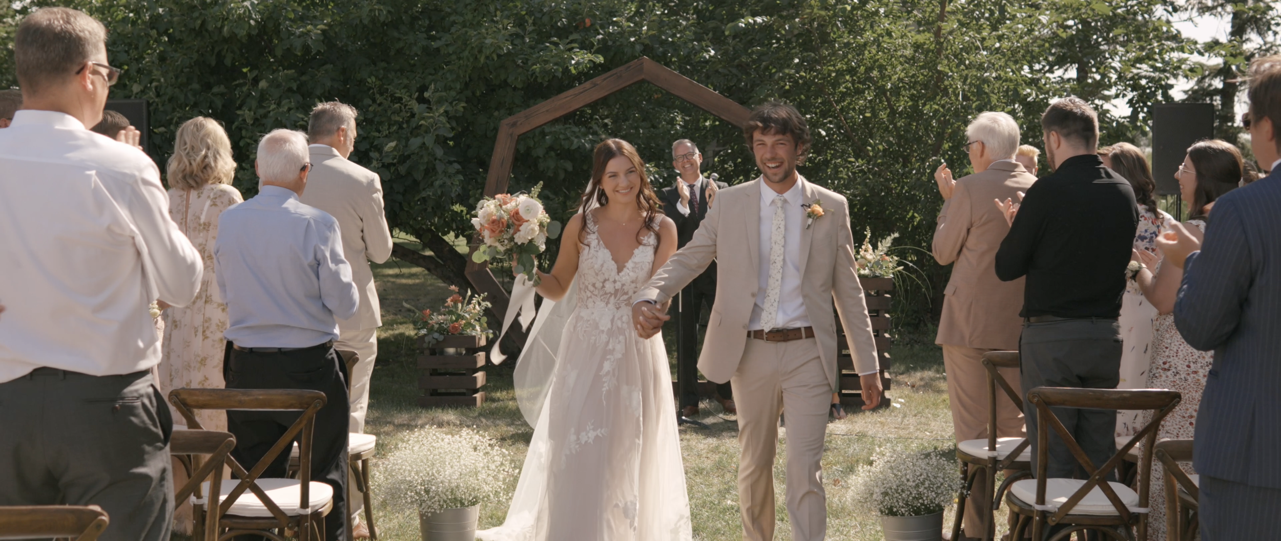 Bride and groom walking down the aisle at an outdoor wedding ceremony, surrounded by guests clapping and smiling, with floral arrangements and a wooden arch in the background.