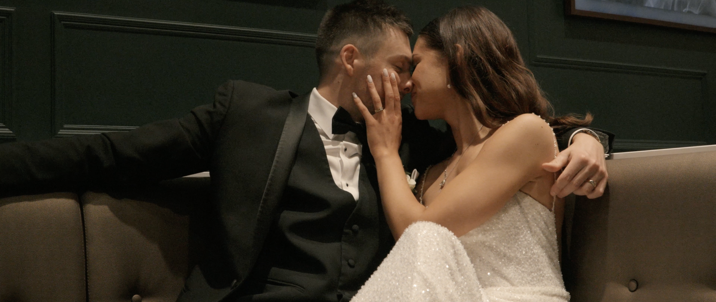 A bride and groom sharing a romantic moment, touching their foreheads and noses together in an intimate pose on a sofa indoors.