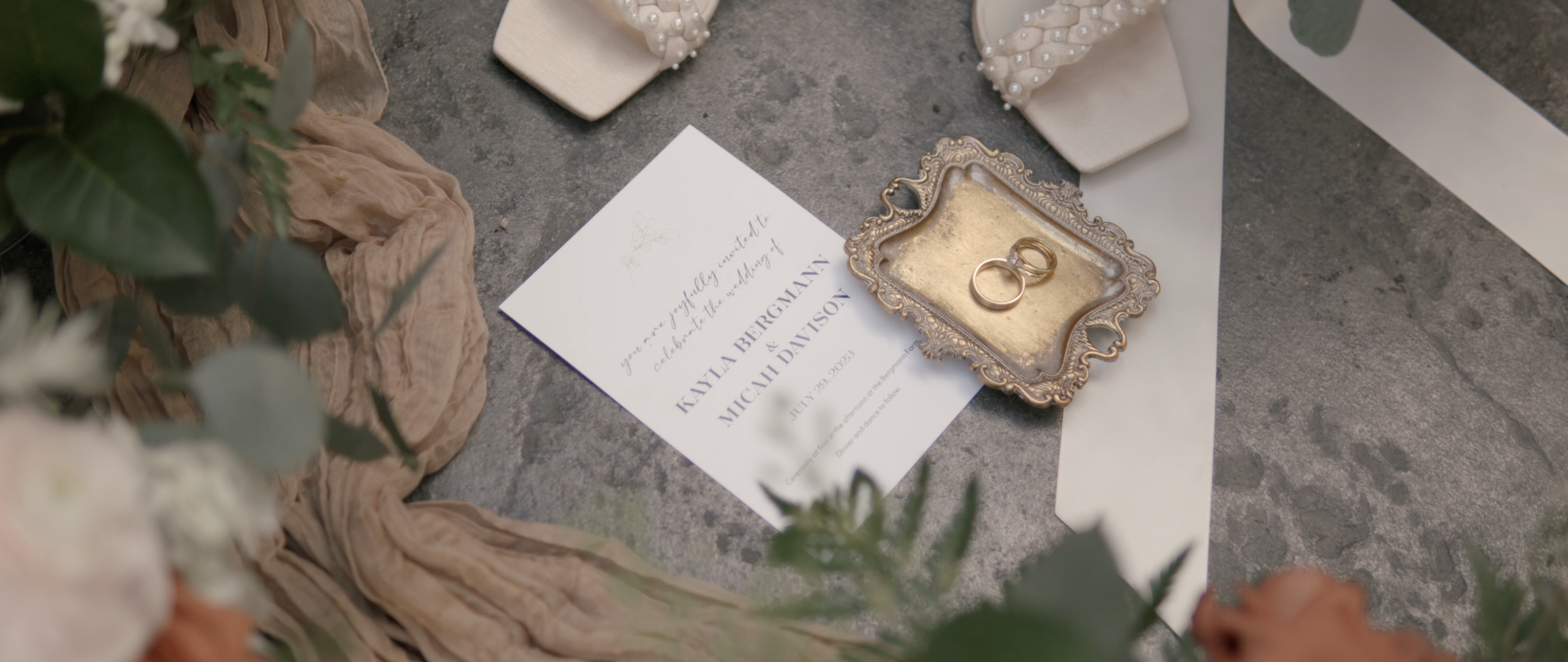 Wedding invitation, two wedding rings on an ornate silver tray, surrounded by flowers, fabric, and ribbon on a concrete surface.