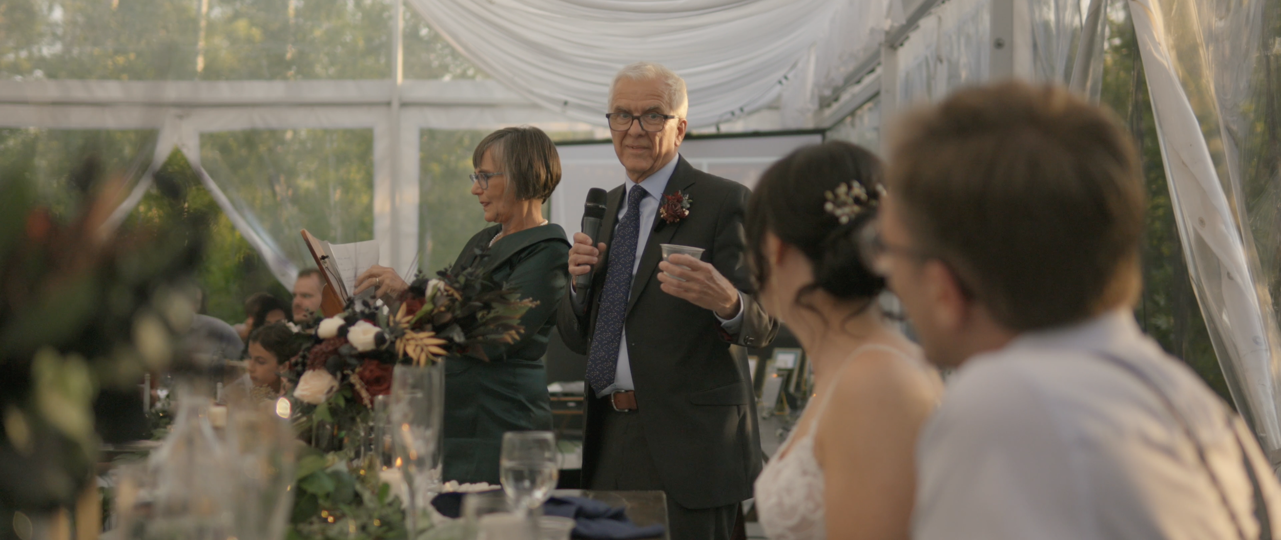 An older man in a suit and glasses is giving a speech with a microphone at a wedding reception held in a glass tent.