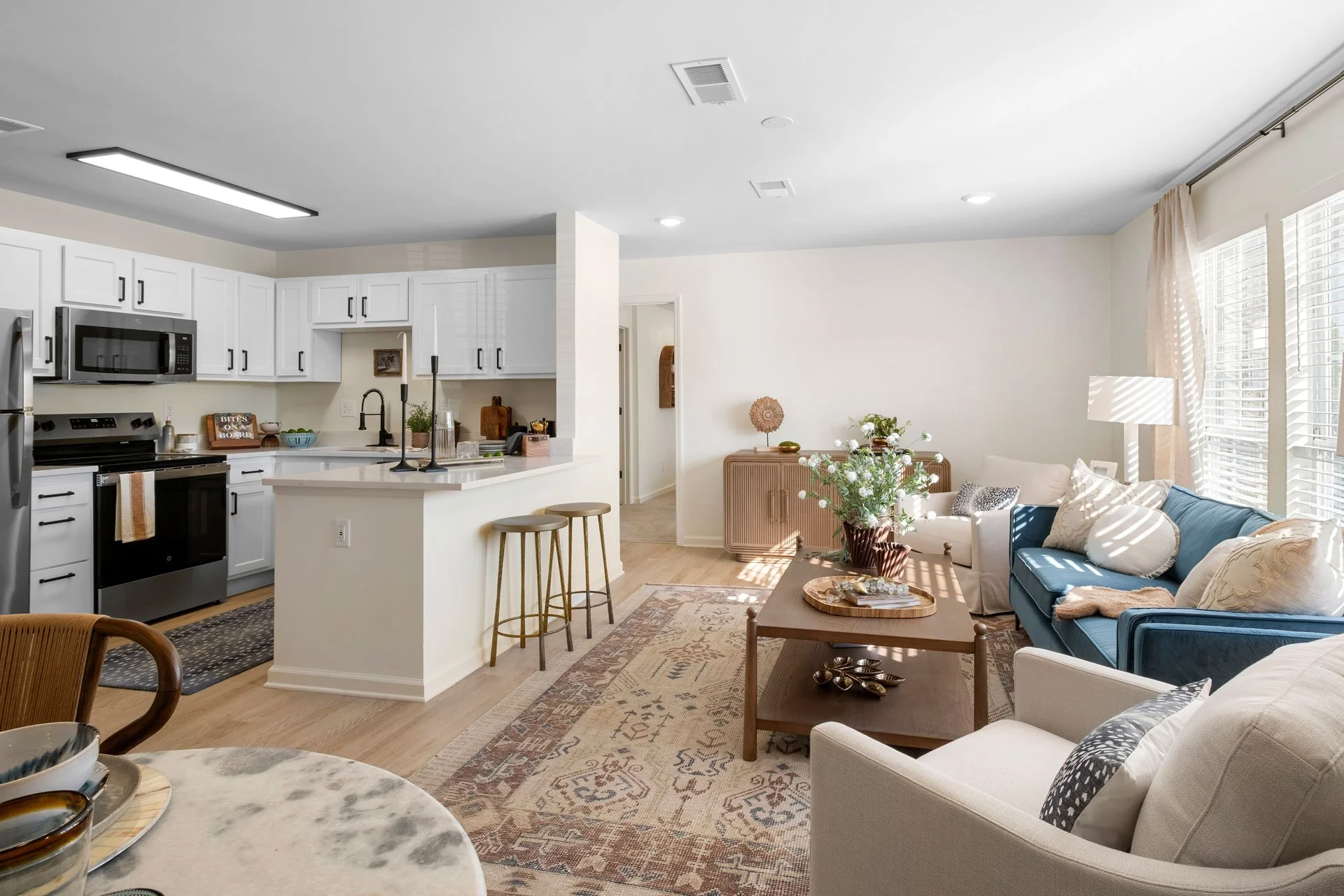 Open-concept living room and kitchen in a modern home, with white cabinets, stainless steel appliances, a blue sofa, and natural light coming through large windows.