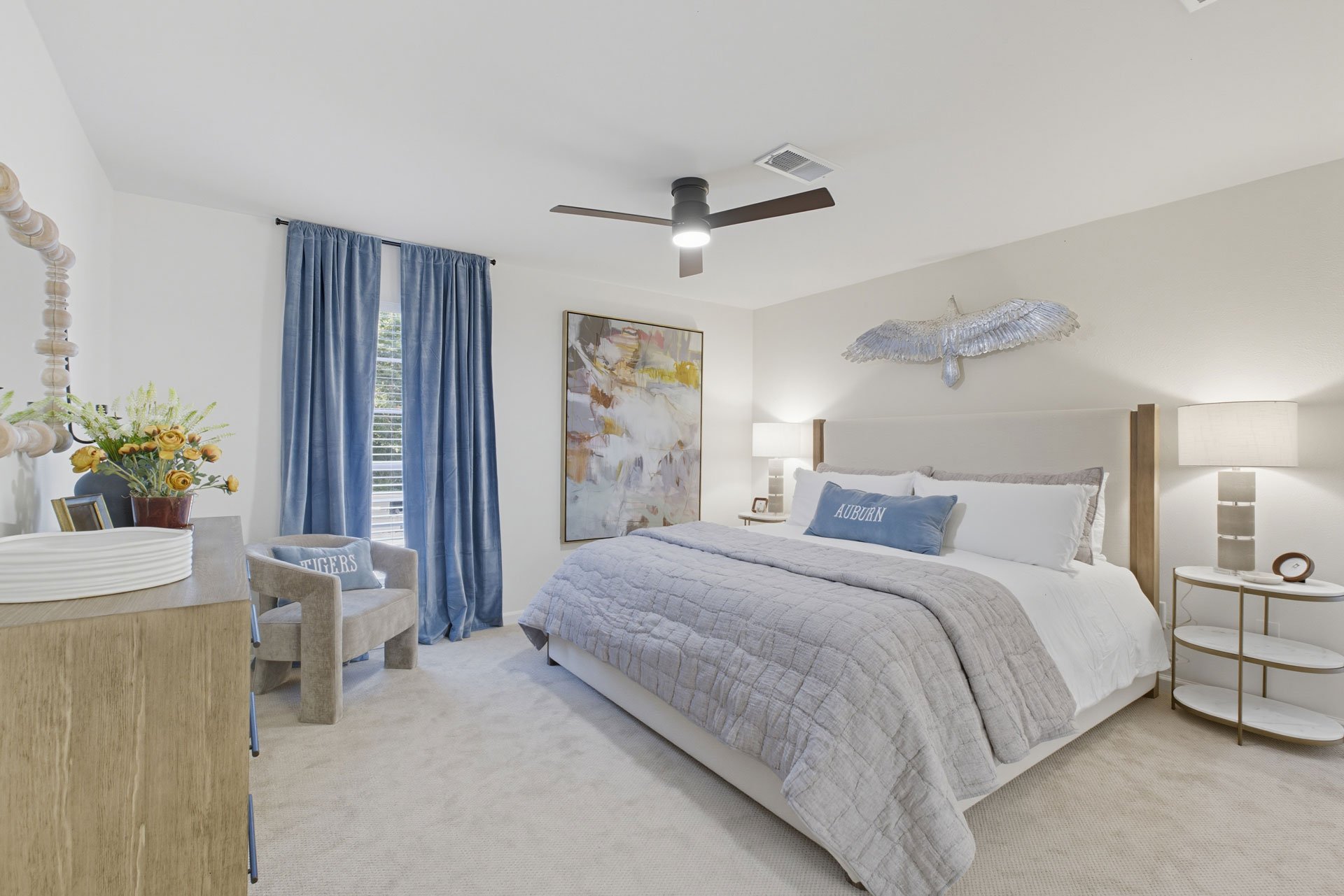 Bedroom with a large bed, decorated with blue and white bedding, blue curtains, a ceiling fan, wall art, a bedside table with lamps, and a wall decoration resembling a bird in flight.