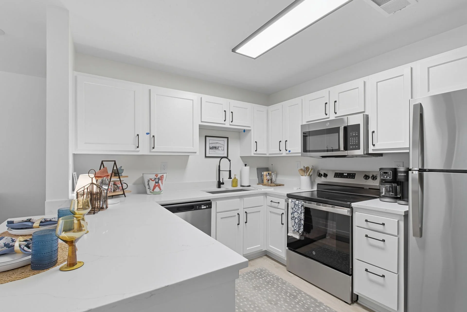 Modern kitchen with white cabinets, stainless steel appliances, a black tap, and a white countertop with tableware and glasses.