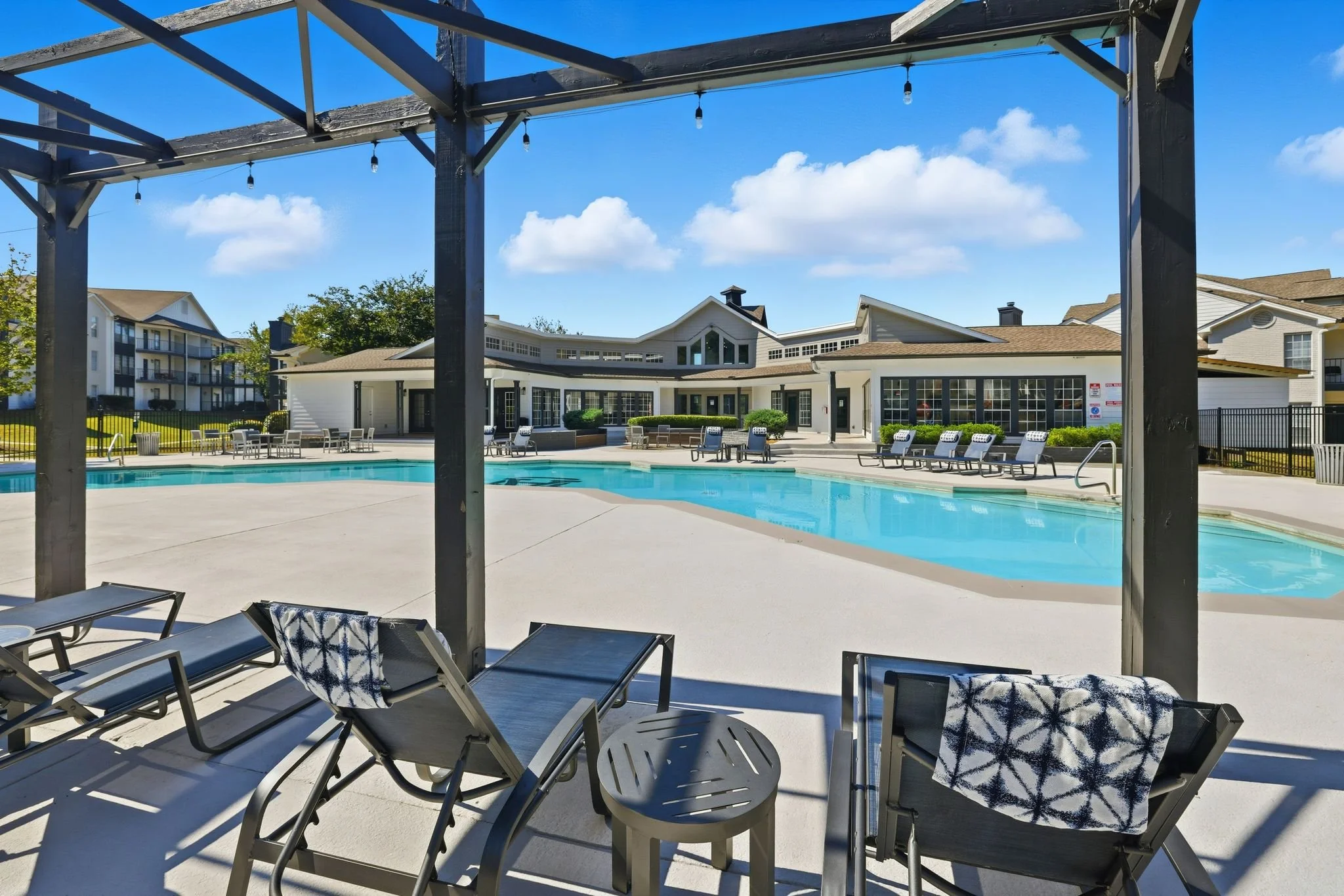 Empty outdoor swimming pool in a residential complex with lounge chairs, umbrellas, and a clubhouse in the background under a bright blue sky.