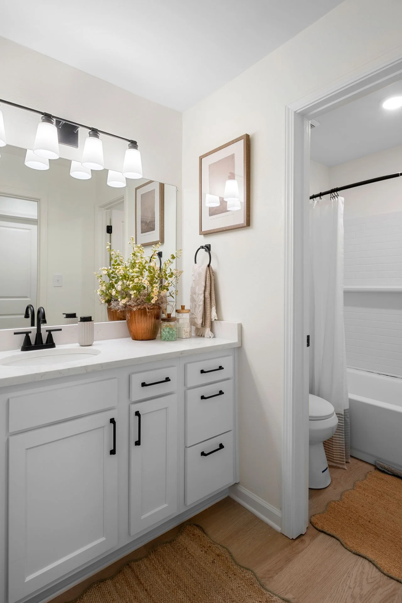 A bathroom with a white vanity, black hardware, a large mirror, a potted plant, and decorative items on the countertop. There is a framed picture on the wall, a towel ring with a towel, and a separate room with a toilet and bathtub/shower visible. Th